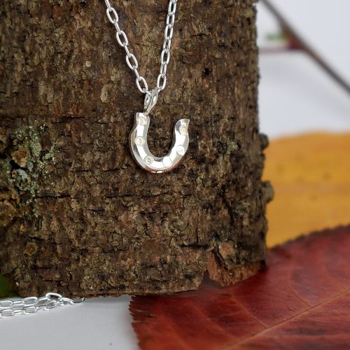 Onnenkenkä-riipus, joka on muotoiltu hopeisen lusikan varresta hevosenkengän muotoiseksi. Pienen riipuksen koko on 9 x 10 mm.