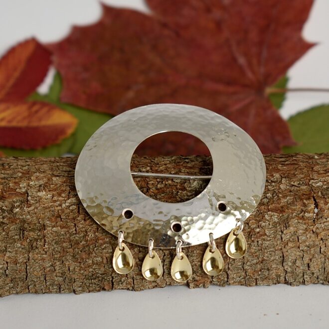 Kuvassa hopeisen jälkiruokalusikan pesästä muotoiltu lappilaistyylinen taottu rintaneula.